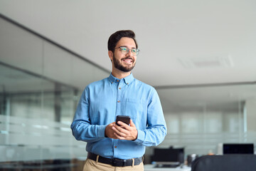 Happy young latin business man holding smartphone standing in office. Smiling hispanic businessman...