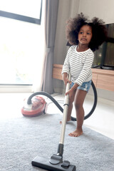 Cute little African girl child with black curly hair vacuuming the floor at bedroom with vacuum cleaner, kid help parents to do housework. Responsible training for children