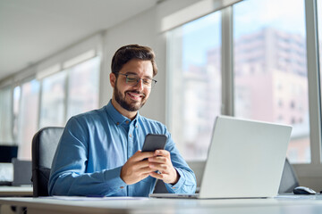 Happy professional latin business man company employee, smiling busy young businessman working on laptop holding smartphone using cellphone looking at mobile phone sitting at office workplace desk.