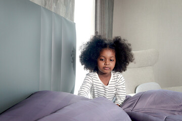 Cute little African girl child with black curly hair making a bed after waking up in morning at...