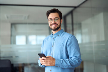 Happy young latin business man holding smartphone standing in office. Smiling hispanic businessman entrepreneur or manager using financial banking apps on cell phone technology device at work. - obrazy, fototapety, plakaty