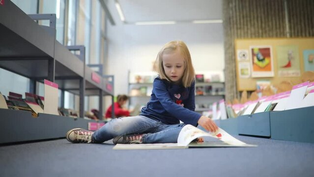 Adorable preschooler girl sitting on the floor in municipal library and reading book