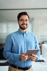Smiling young latin business man manager using tablet computer, happy hispanic businessman employee, company worker looking at camera holding tab working standing in office. Vertical portrait.