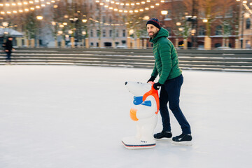 Joyful man skating with a bear-shaped aid on a festive ice rink in the evening