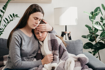 Caring mum comforting sad ill kid, expressing love and support, child psychologist concept. Young caucasian mother hugging sick upset little daughter covered blanket, sitting together on sofa at home