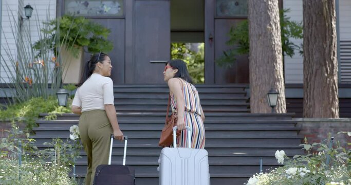 Happy asian female friends with suitcases walking on stairs of home on sunny day, slow motion