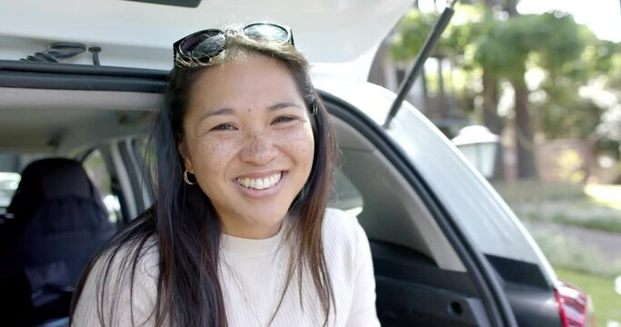 Happy asian woman with sunglasses sitting in car trunk and smiling on sunny day, slow motion
