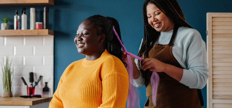 Happy African American hairdresser braiding hair to a female customer in salon