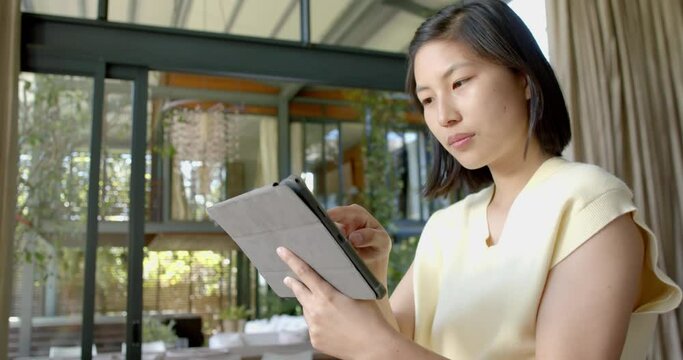 Happy asian woman standing and using tablet in sunny living room, slow motion