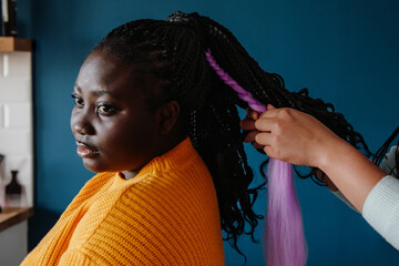 Close-up of hairdresser braiding hair to young African woman in salon - obrazy, fototapety, plakaty