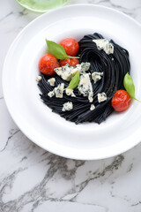 Plate with black spaghetti, blue cheese, roasted tomatoes and basil, vertical shot on a white marble background, elevated view