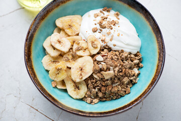 Turquoise bowl with granola, banana chips and white yogurt, horizontal shot on a white granite background, middle closeup