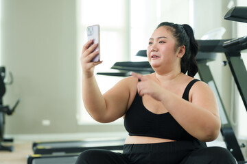 Chubby  woman sitting and resting after exercising in fitness center. Video call with friends via...