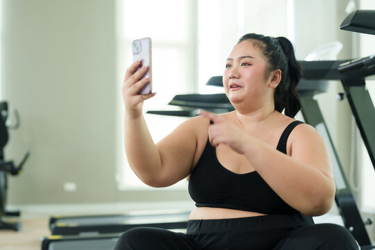 Chubby  woman sitting and resting after exercising in fitness center. Video call with friends via smartphone To invite you to exercise The joy of a cute fat Asian woman trying to lose weight