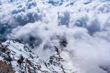 alpine landscape with peaks covered by snow and clouds Cloudy mountains. Mountains in clouds at sunrise in summer. Aerial view of mountain peak with green trees in fog. Top view o