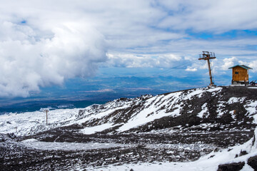 Mount Etna national park in winter. View from volcano crater with black volcanic lava stones and snow under cloudy sky and smoke. Cable car and view over Sicily island, Italy