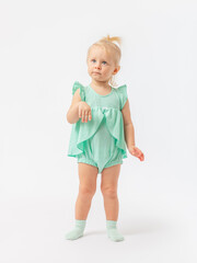 Arrogance, resentment. A 2-year-old girl with an expressive serious look on a white background in a green bodysuit dress.