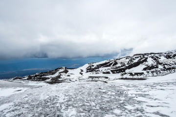 Fototapeta na wymiar Mount Etna national park in winter. View from volcano crater with black volcanic lava stones and snow under cloudy sky and smoke. View over Sicily island, Italy