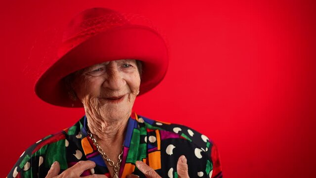 Closeup funny view of happy elderly toothless senior woman wearing red hat pointing fingers herself ask say who me, smiling talking and pointing. Isolated on red background in slow motion