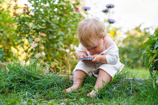 Little child baby in a muslin white suit in the garden at the dacha in summer playing with a smartphone, happy active summer