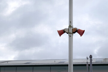 Two red, slightly rusty bullhorn claxons are clamped to a white metal pole. An indsutrial unit can be seen in the background.
