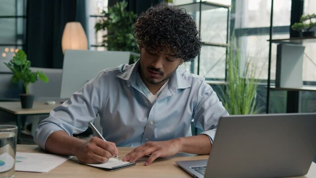 Focused businessman business person accountant office worker writing ideas in notebook paperwork Arabian Indian man student write notes data information e-learning laptop computer educational course
