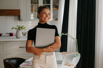 Attractive young woman holding laptop and smiling while standing at home office
