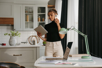 Concentrated young woman talking on mobile phone and using digital tablet while working at home