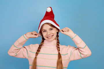 Obraz premium Little smiling girl wearing and holding Santa hat on blue background.