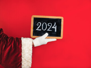 Santa Claus's hand in a white glove holds a board with the New Year 2024 number written in chalk.