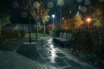 Night park paths and colored houses on a rainy autumn night. Night paths, benches, lanterns in a...