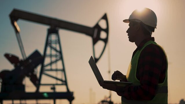 Oil industry. Engineer worker makes notes on operation of an oil pump. The concept of mining. An engineer with laptop and a tablet checks operation of an oil pump. Industrial production of crude oil