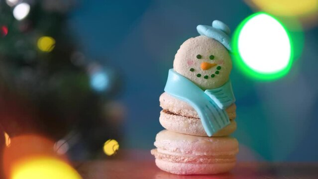 Snowman dessert macaron decorated with hat and scarf with colorful lights and bokeh flashing on foreground and Christmas tree on background. Merry Christmas and Happy New Year festive concept