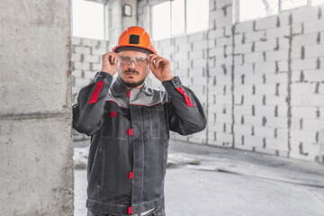 Construction worker in a hard hat and overalls adjusts his safety glasses at a construction site,...
