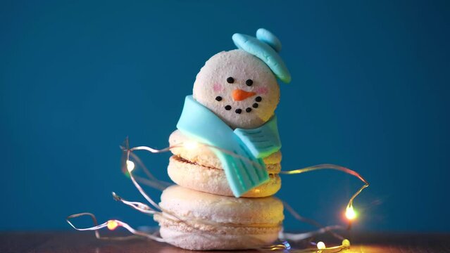 Snowman dessert macaron decorated with hat and scarf wrapped with colorful decorative flashing garland lights against blue background. Camera moves away from object. Merry Christmas concept