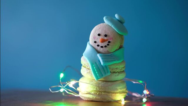Snowman dessert macaron decorated with hat and scarf wrapped with colorful decorative flashing garland lights against blue background. Camera moves away from object. Merry Christmas and HNY concept