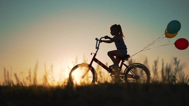 Dream kid. Silhouette of kid on bike in park. Girl rides in park on green grass. Child games in nature.Traveling with balloons on bike.Active child freedom in summer.Girl learns to ride bike in nature
