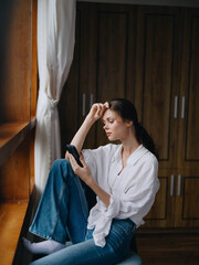 Young woman sitting at the window on a chair with a phone in hand smiling and looking out the window, spring mood, home cozy atmosphere, aesthetic lifestyle.