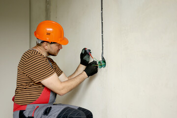 Electrician male in uniform, protective gloves and helmet checks presence of electrical voltage in...