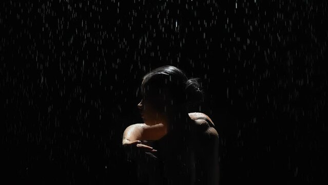 Silhouette elegant female dancing posing under rain at night. Seductive woman, gymnast dancer wearing black swimsuit, raindrops splashes, water drops roll down sexy body in shower, black background