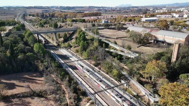 Trenes Renfe R2 Rodalies entrando y saliendo de la estaci&oacute;n de Santa Perp&egrave;tua de Mogoda, con vistas a Montserrat y al lago de Parc Central y un partido de f&uacute;tbol, grabado desde dron 4k 60fps.