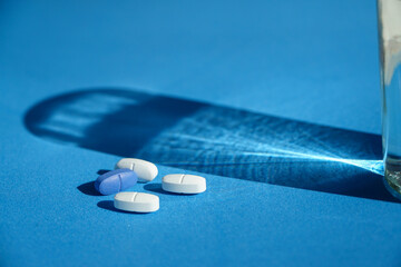 Close-up view of pills on a sky-blue surface with a glass of water and its shadow.
