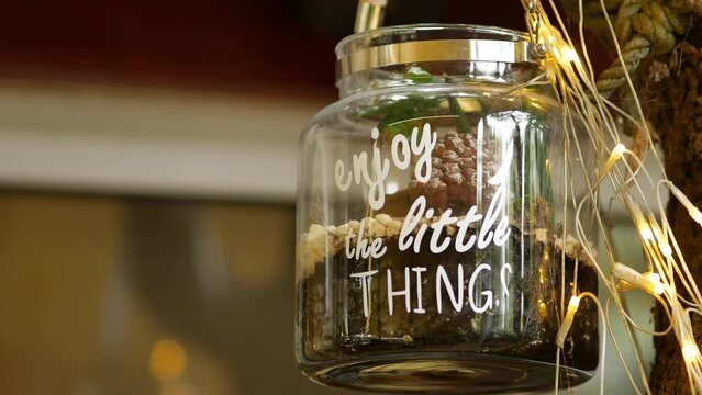 Glass jar with text quote Enjoy the Little Things hanging on a white rope on a tree branch. LED light garland. Halloween decoration to attract guests to restaurant. Rovinj, Croatia, November 14, 2023
