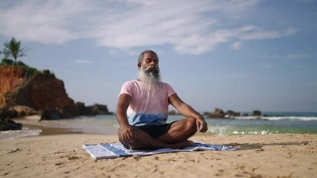 Old man meditating and sitting in lotus pose to sound of waves on paradise island beach at ocean shore. Black male senior experiences zen state of mind in yoga at exotic coast with crossed legs.