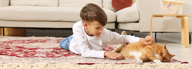 Little boy petting cute ginger cat on carpet at home. Banner design © New Africa