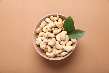 Tasty cashew nuts and green leaves in bowl on pale brown background, top view