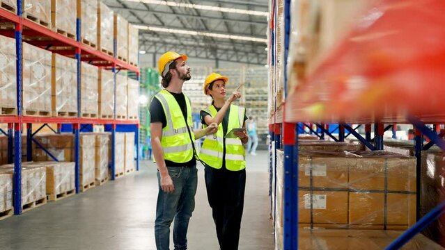 Retail Storehouse full of shelves with goods in cardboard boxes, Young man and woman warehouse worker wearing vest and helmet safety discuss product delivery