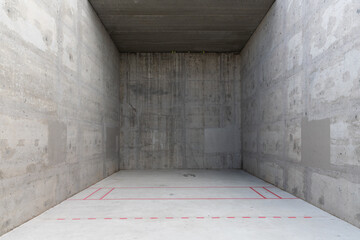 A concrete racquetball court with red markings on the ground at a public park. The bare cement walls have a grungy look and a splotchy, unfinished texture. The end of the court is cast in shadow.