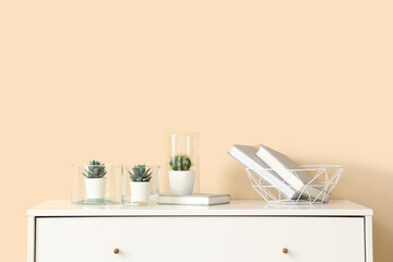 Different houseplants and books on chest of drawers near beige wall