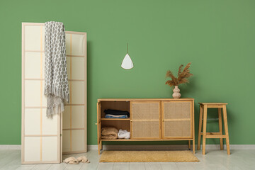 Modern light wooden chest of drawers with folding screen and stool near green wall in room